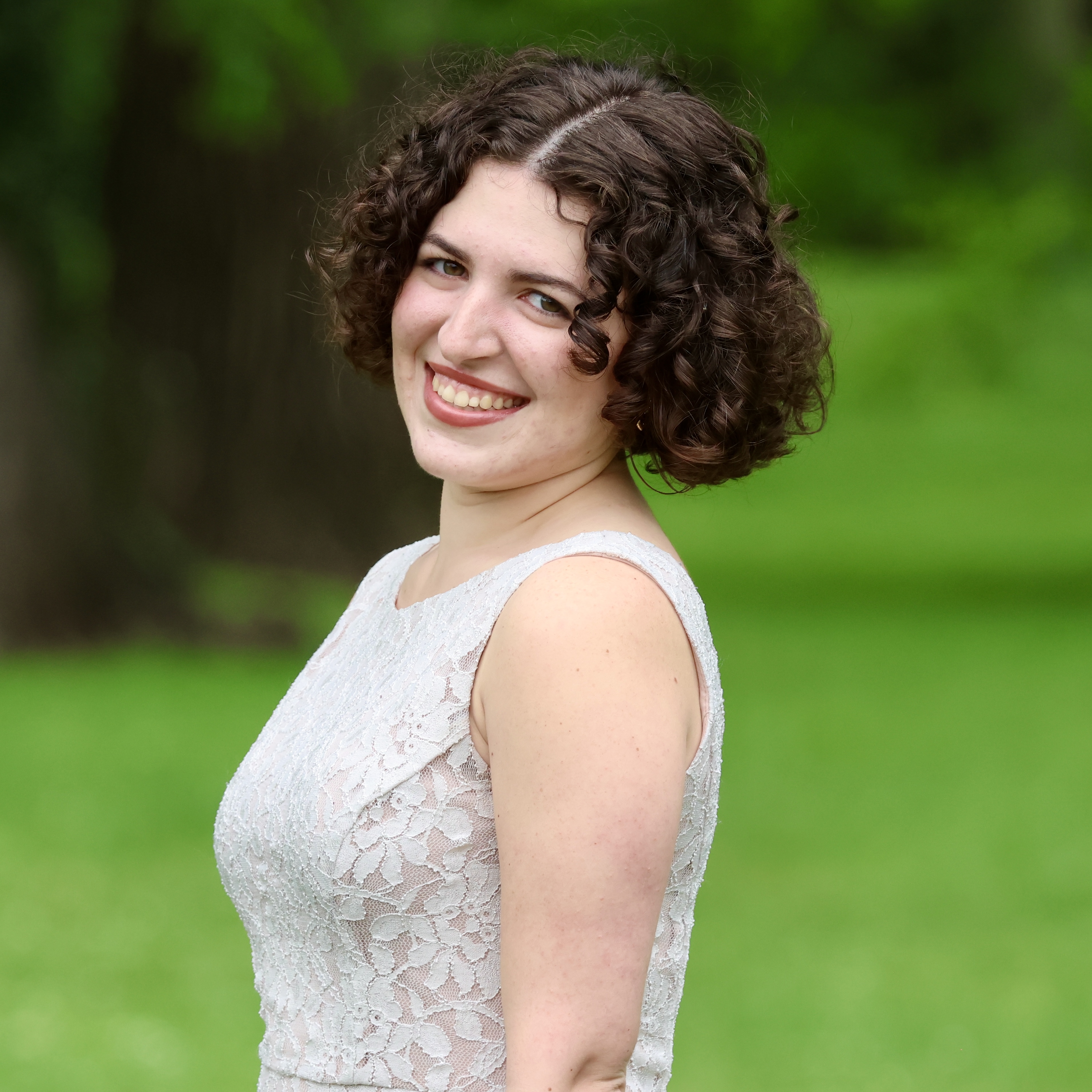 A young woman with curly brown hair, wearing a white lace sleeveless dress, posing for a photo in a grassy field with tree...